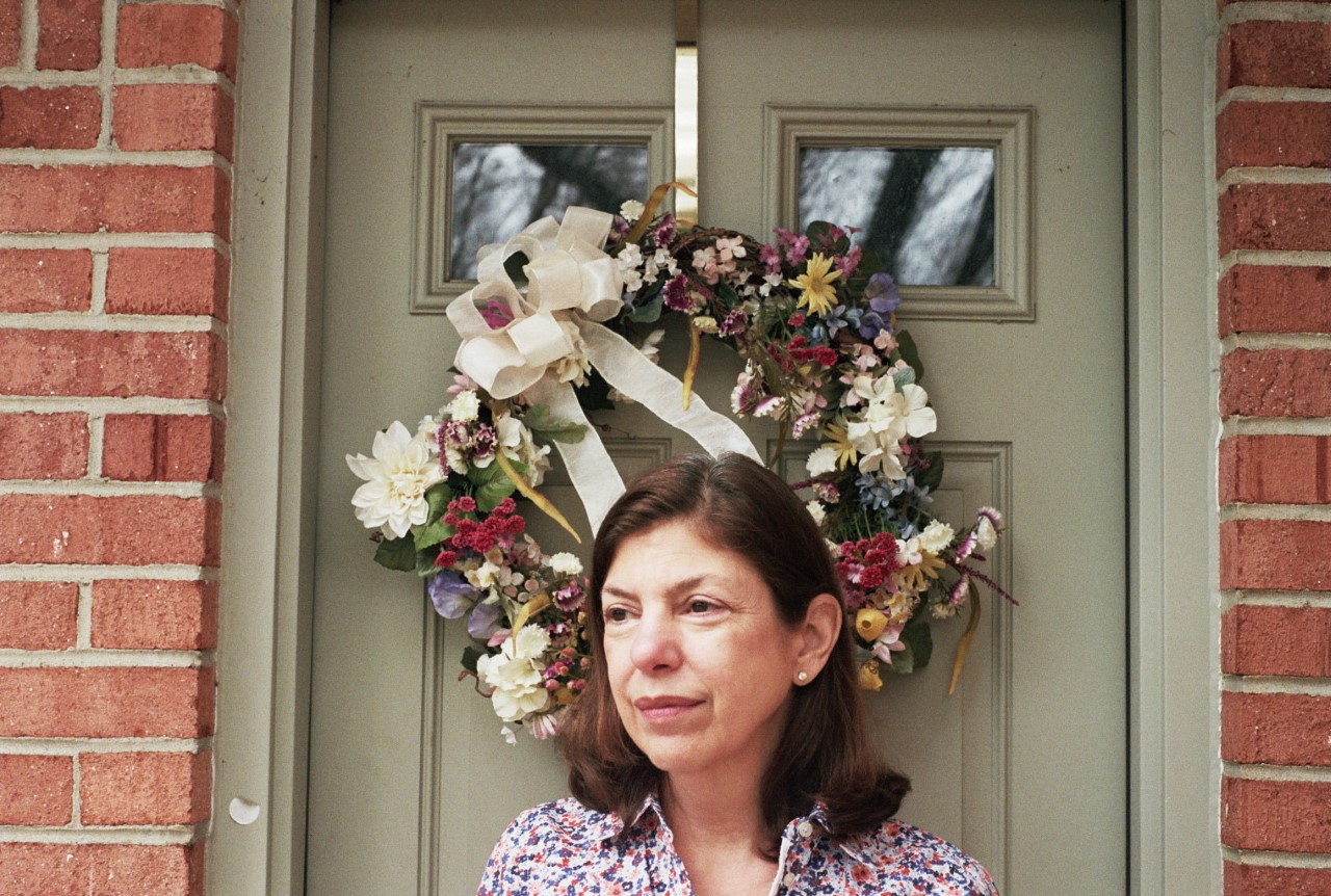 Elizabeth has shoulder length brown hair and is wearing a floral collared shirt. She is standing in front of a door with a floral wreath.