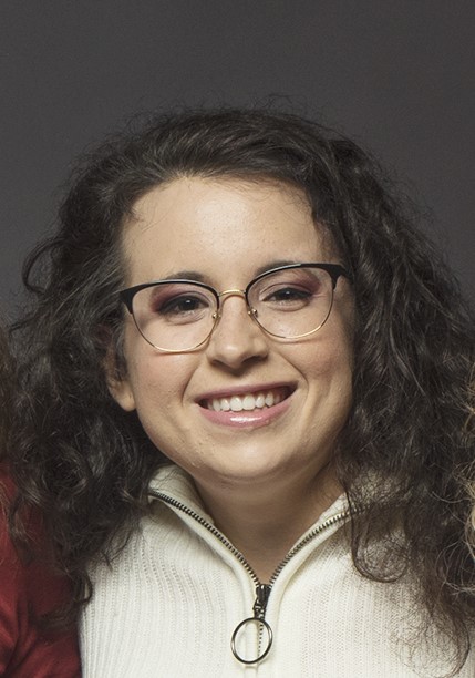 A picture of a young woman smiling. She is wearing a white zip-up fleece and glasses. She has long curly brown hair.