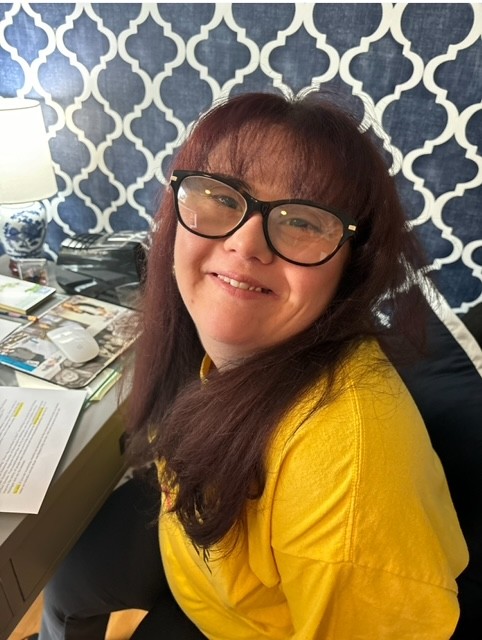 A picture of Allie Hayes. She is a young woman with long brown haor, glasses, and is wearing a yellow long-sleeved shirt and smiling.