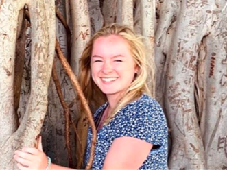 Sam is standing by a big tree. She is wearing a blue and white shirt, is smiling, and has shoulder-length blonde hair.