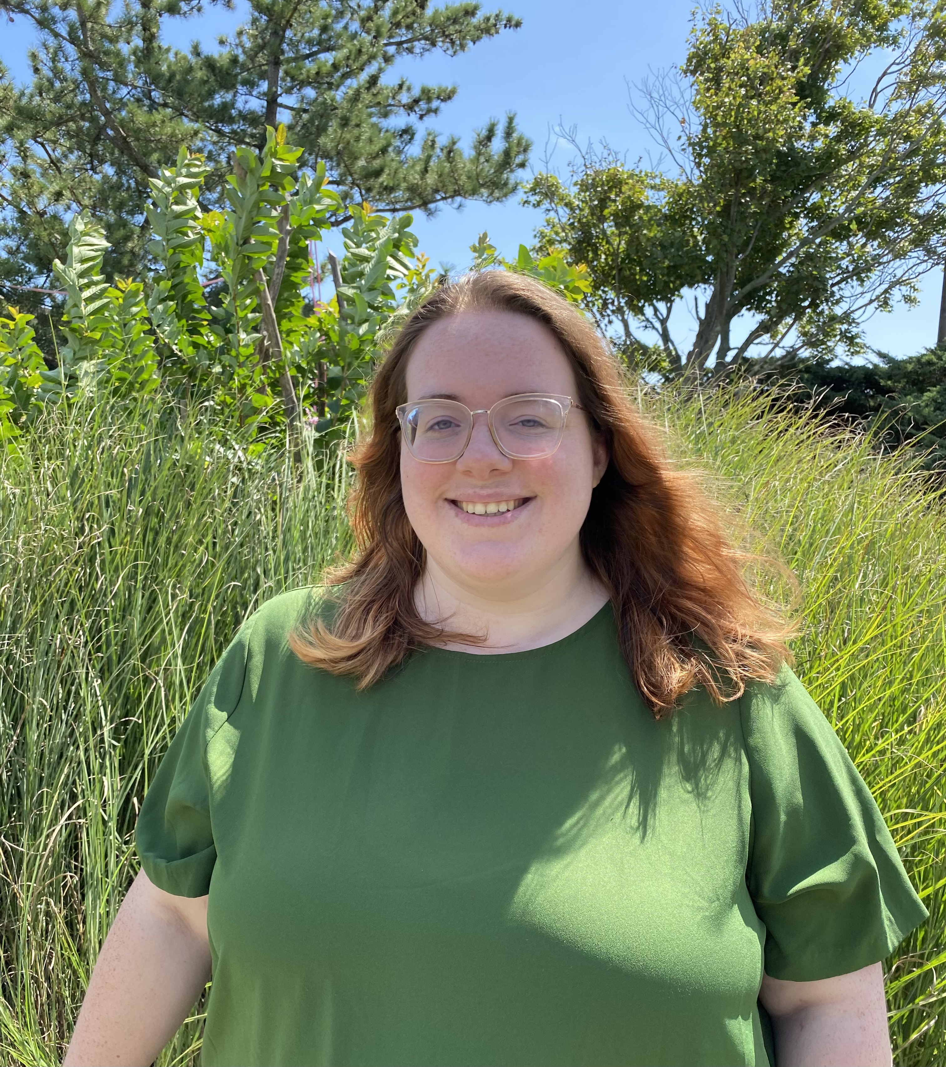 Laura is standing outside in a field. She has shoulder-length auburn hair, is smiling, wearing glass and a green shirt.