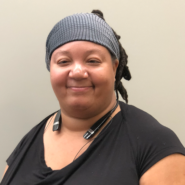 Picture of a black women who is smiling. She is wearing a grey and black head scarf, and a black shirt.