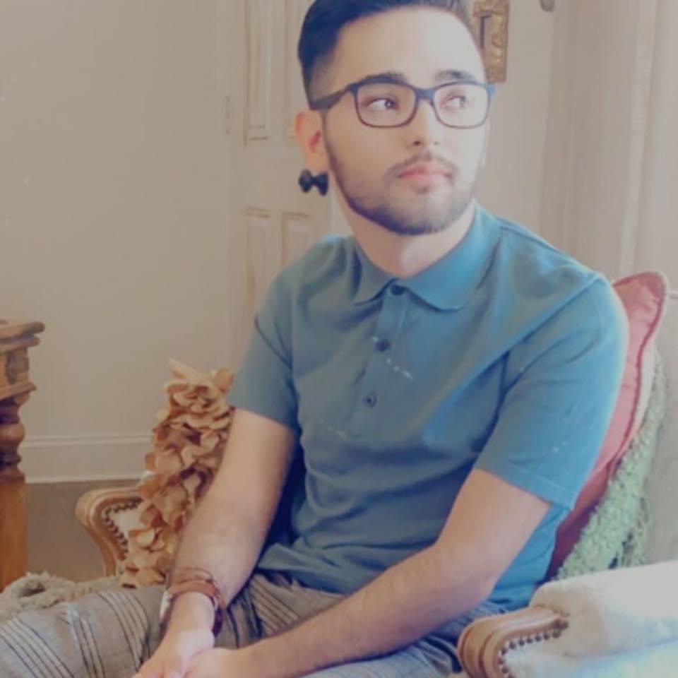 Picture of Leonardo Bravo. Leo is sitting in a chair, wearing a blue shirt, striped pants, and black glasses. He has short black hair.