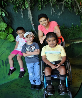 A mother and her 3 children posing in front of a wall with vines and leaves at the Detroit Zoo. The young boy in front is in a wheelchair.