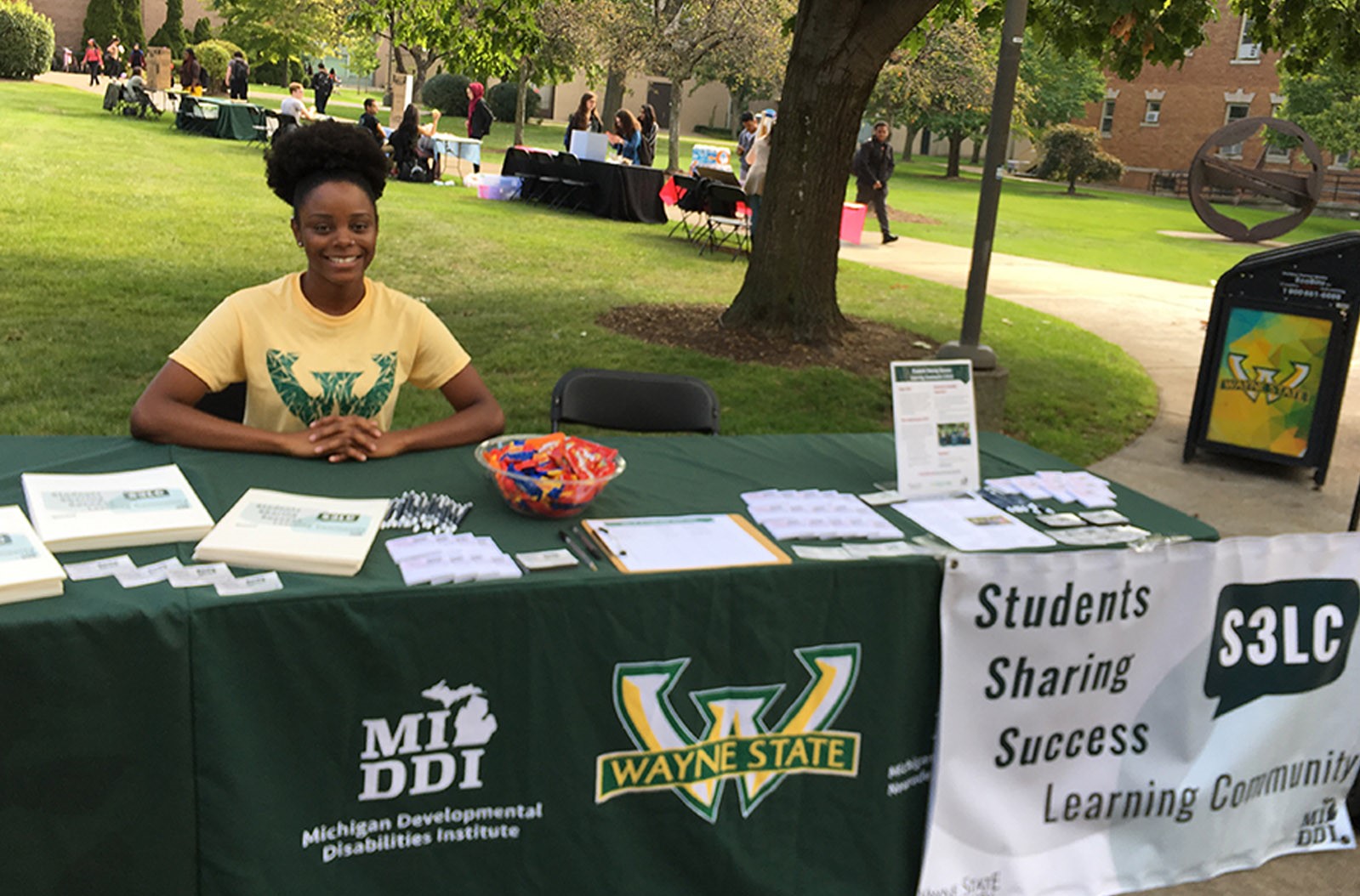 A picture of Nia Anderson sitting at a long table outside on a lawn on the Wayne State campus. Nia has curly black hair that is up and she is wearing a yellow and green Wayne State t-shirt.