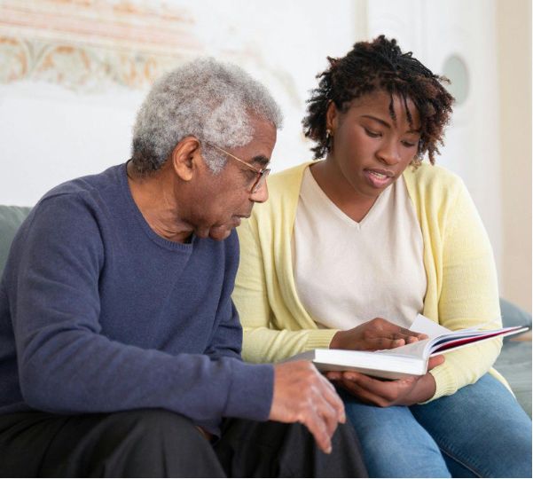 A younger person helping an older person with reading. Both are looking down at the book while seated on a couch.