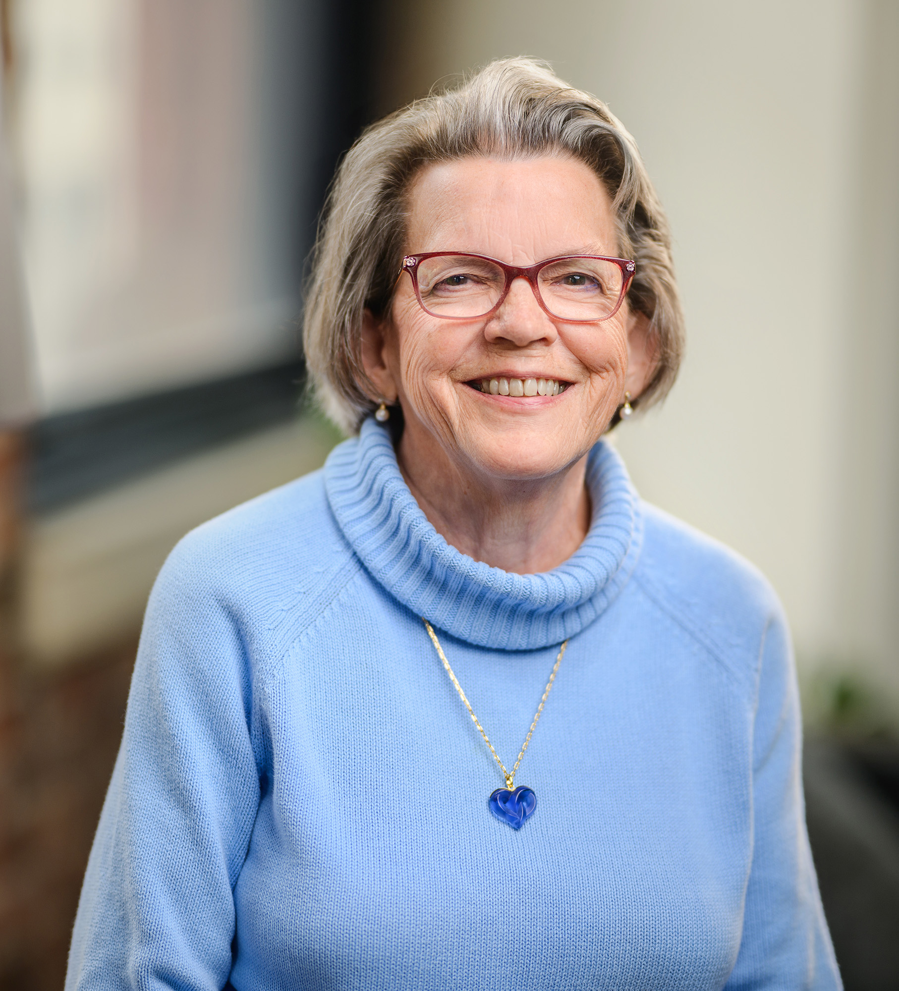 Photo of Barbara LeRoy. Barb is wearing a blue sweater and a necklace with a blue heart pendant. She has shoulder length gray hair, reddish brown glasses, and is smiling.